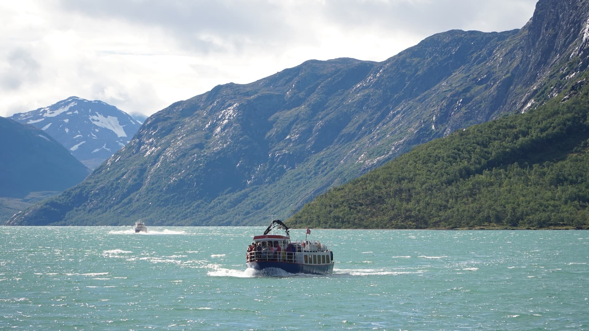 Ferry boat on Gjende lake with steep, forested mountains and snow-capped peaks.