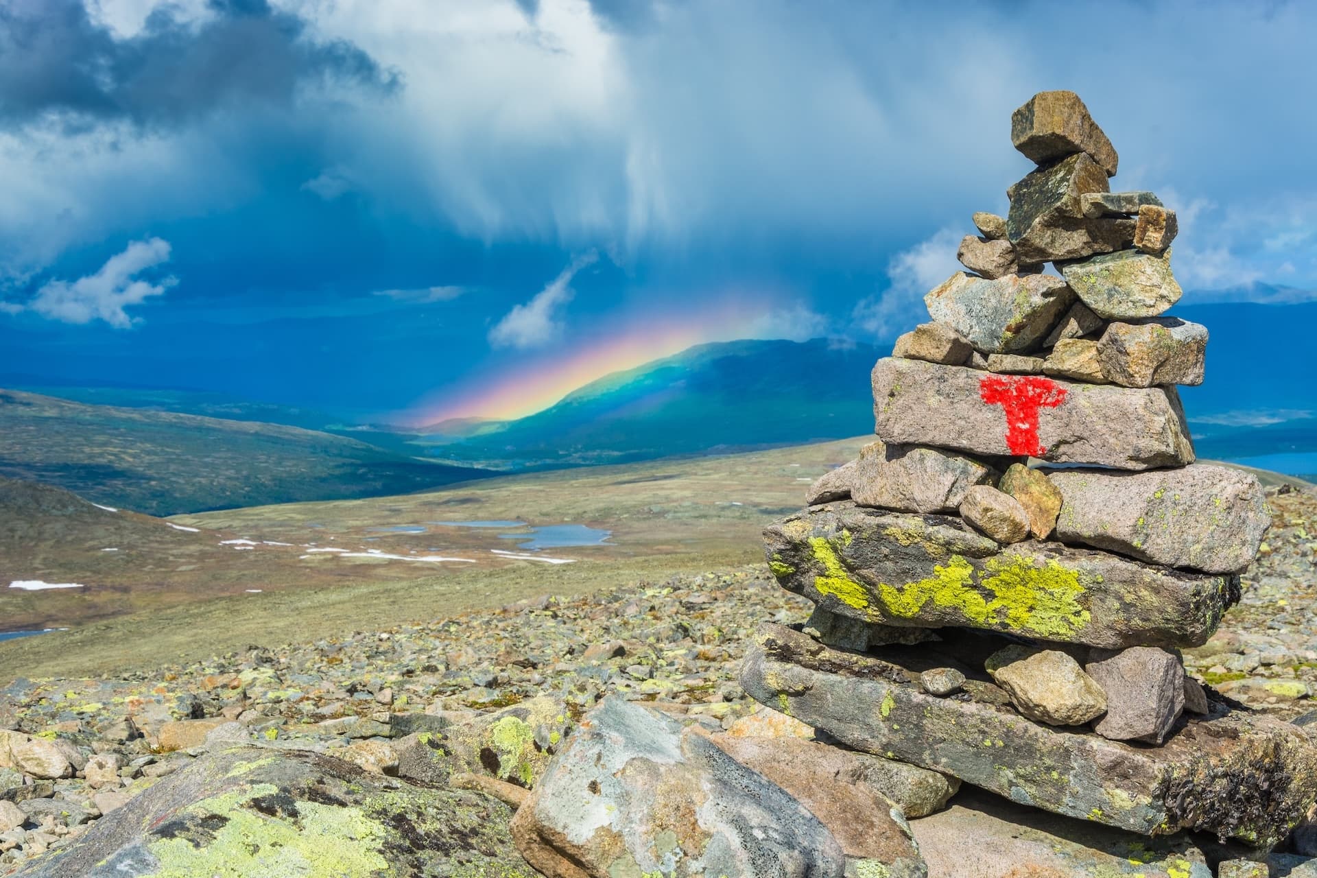 Hiking cairn with red T-mark overlooking mountain valley with rainbow and dramatic sky in Norway.