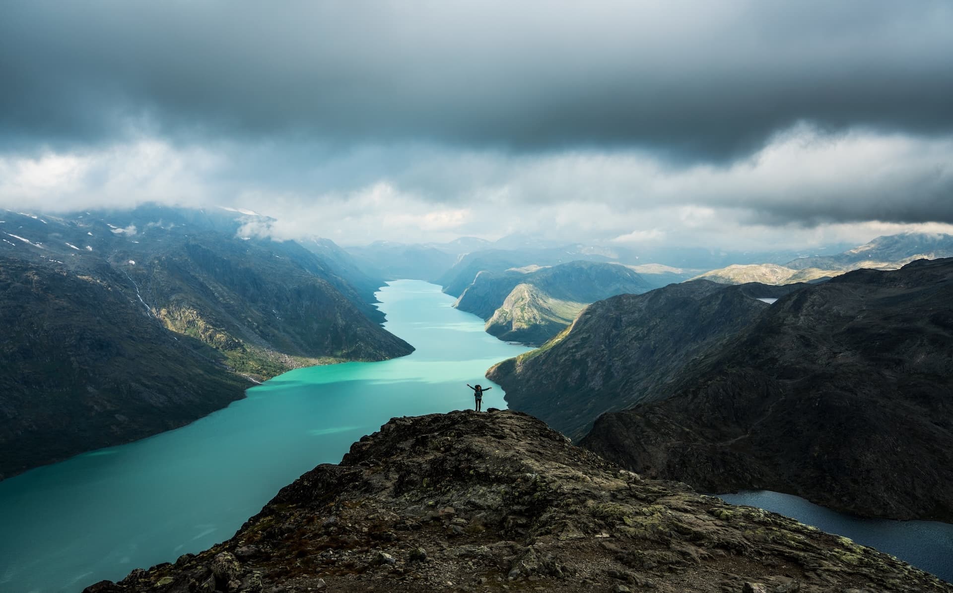 Hiker on ridge overlooking turquoise glacial lake surrounded by mountains under dramatic clouds, Besseggen.