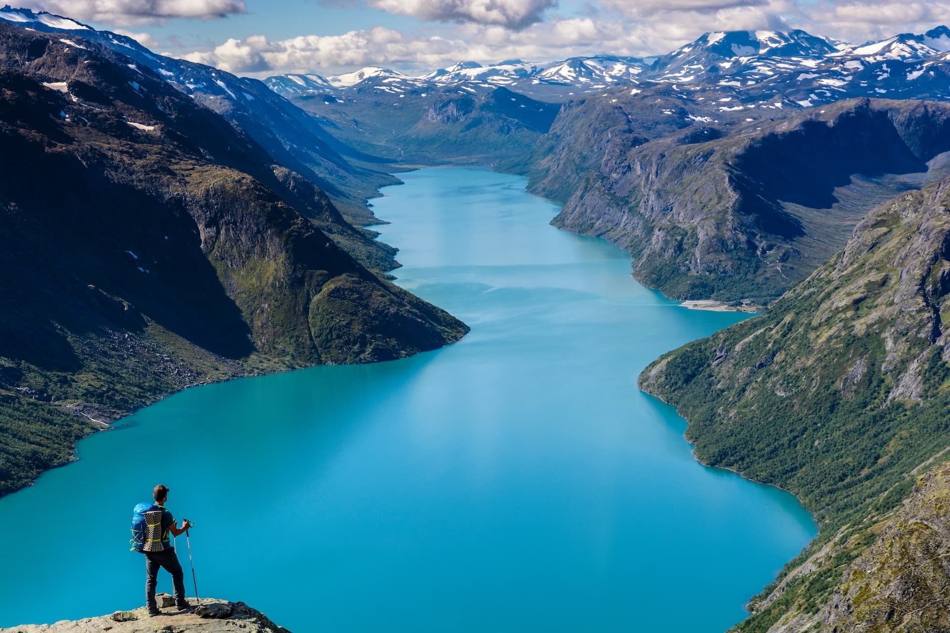 Hiker overlooking bright turquoise glacial lake surrounded by steep, rocky mountains with snow patches.