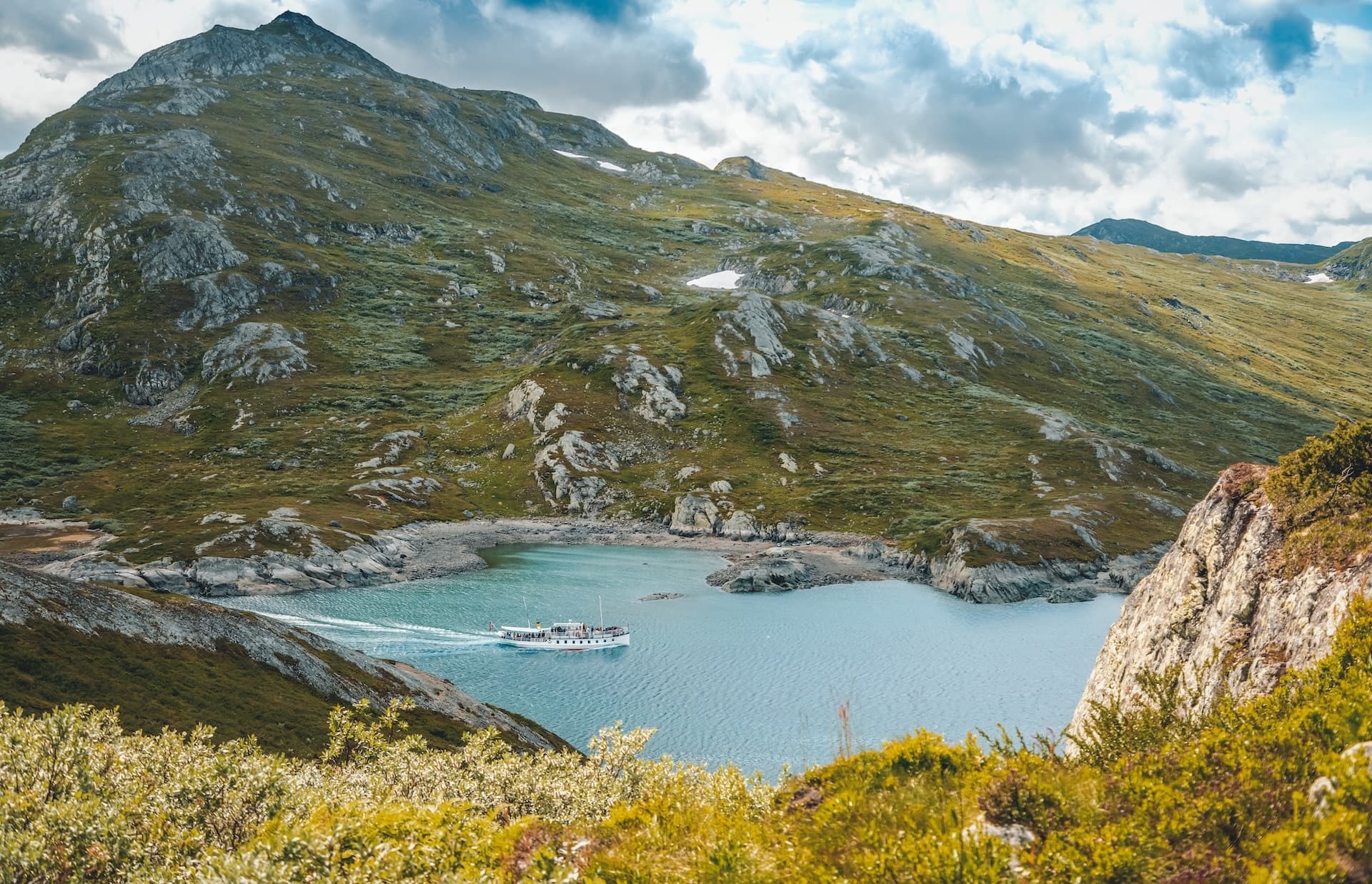 Ferry on the Gjende lake in Norway surrounded by green, rocky mountains under a cloudy sky.