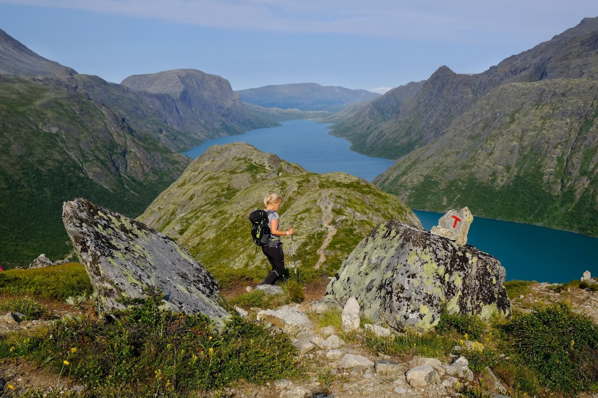 Hiker on Gjendesheim trail with trekking poles overlooking steep mountains and blue lake.