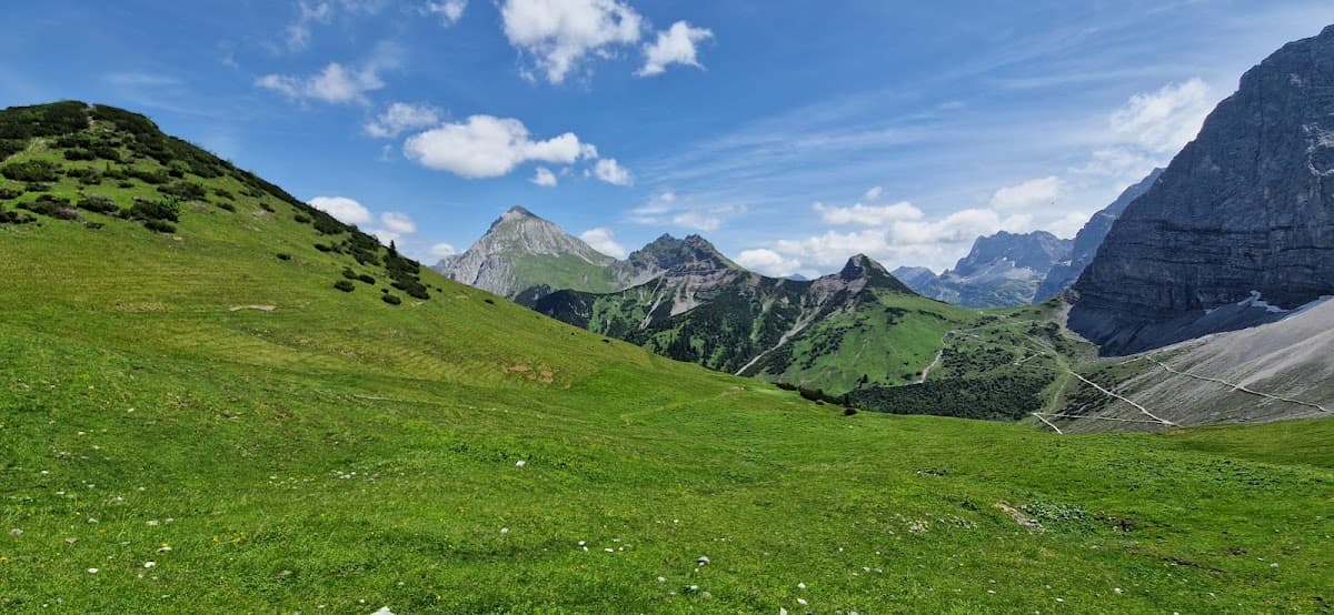 Vast green alpine meadow sloping toward rugged mountains under a blue sky with white clouds.