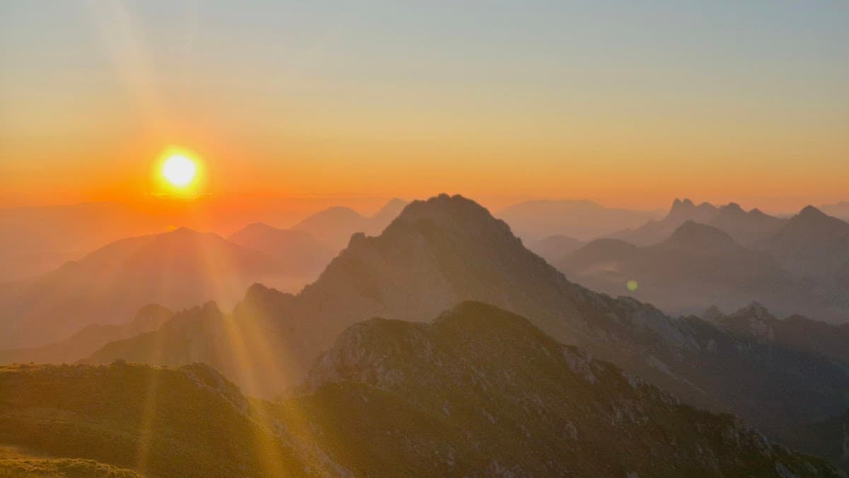 Mountain range at sunrise with sun flare over hazy peaks and foreground ridge.