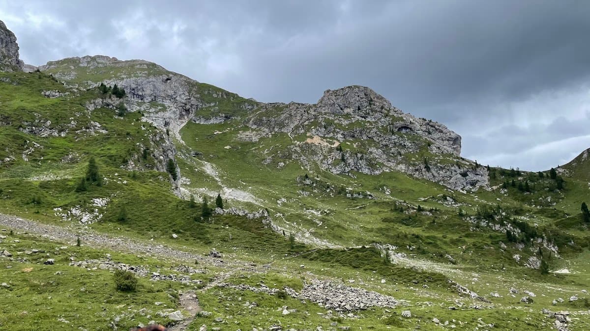 Grassy mountain slopes with rocky outcrops under a cloudy sky, hiking trail visible.