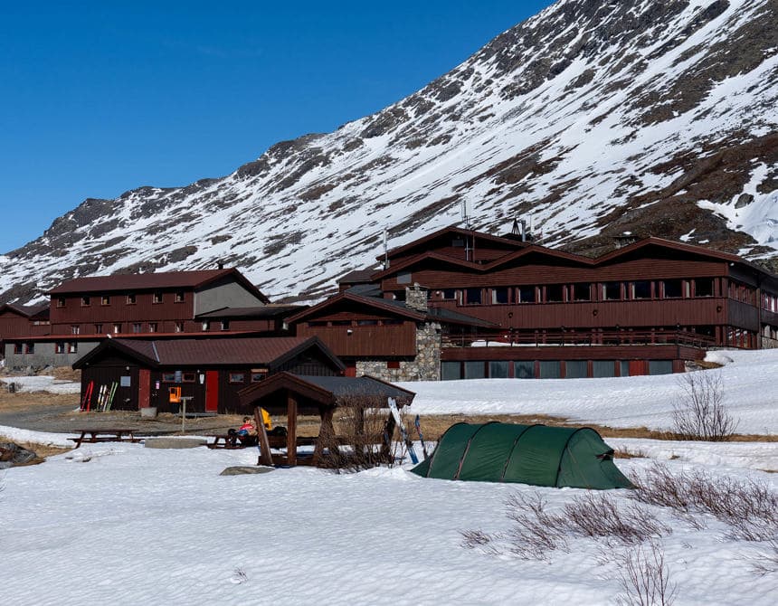 Green tent pitched in snow near wooden mountain lodge below steep, snow-covered slope.