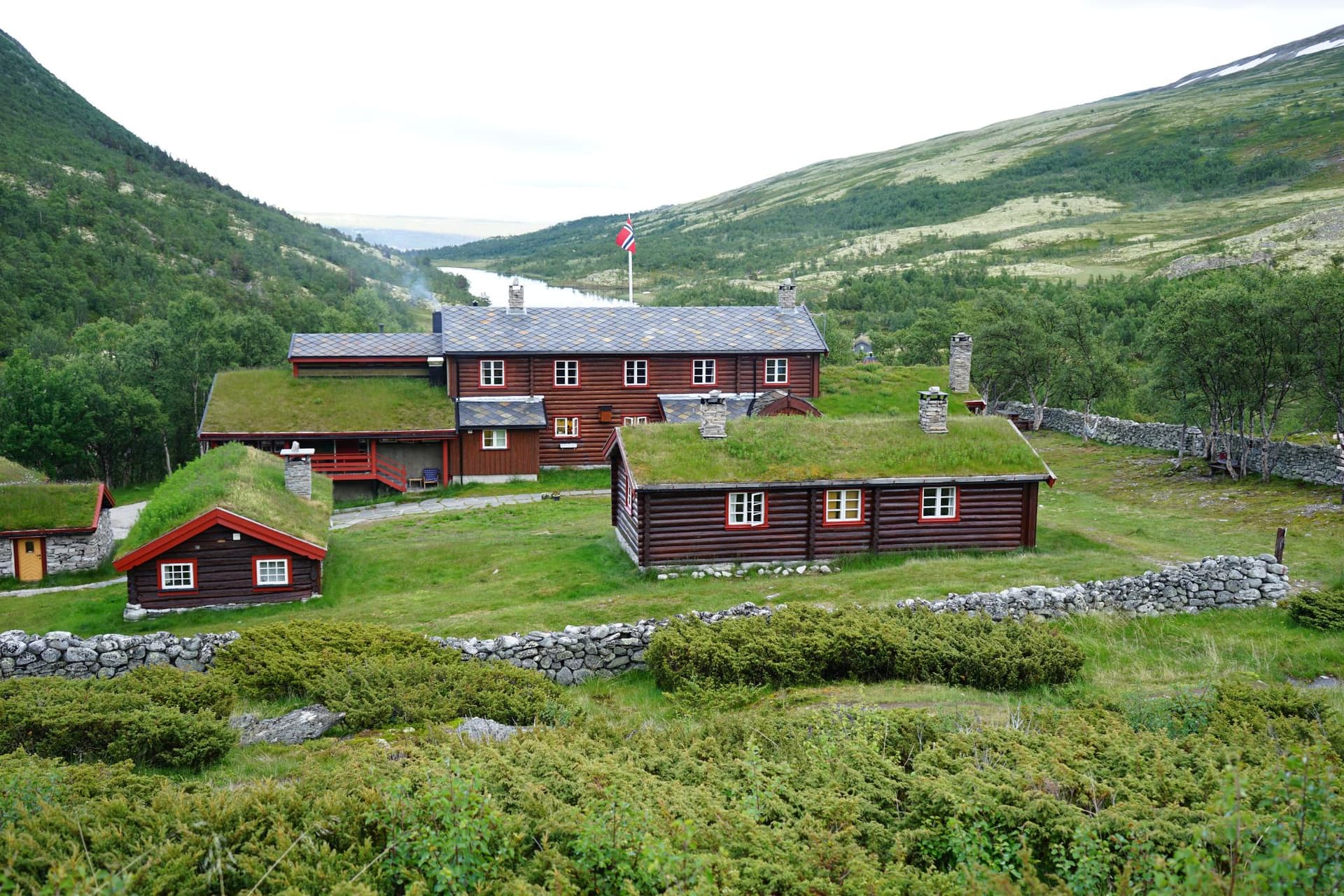 Log cabins with grass roofs and a Norwegian flag near a mountain lake valley