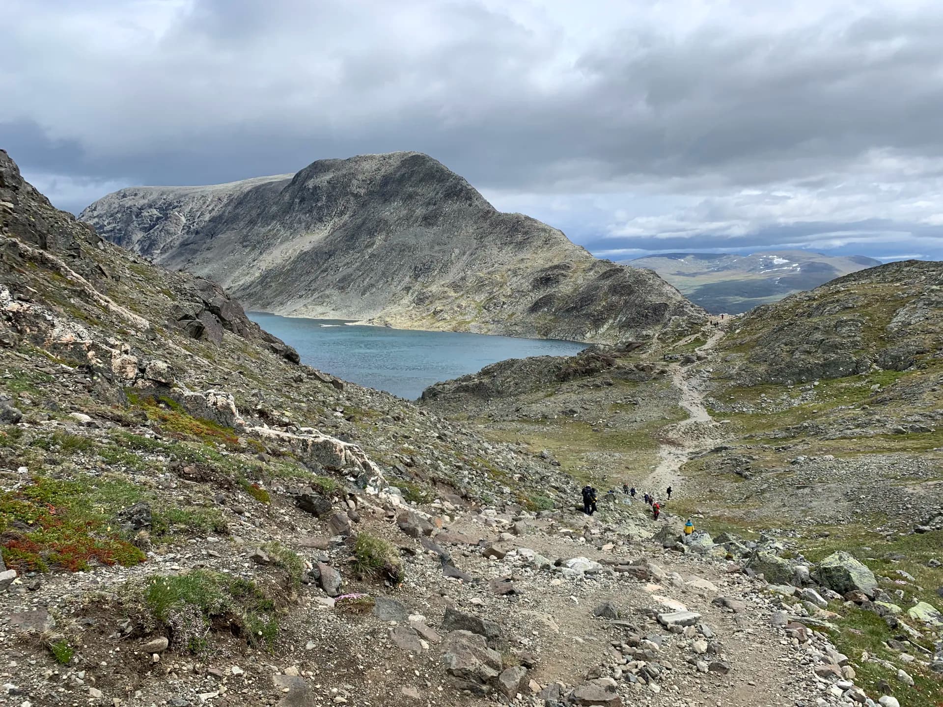 Hikers on a rocky mountain trail overlooking a blue alpine lake in Jotunheimen, Norway.