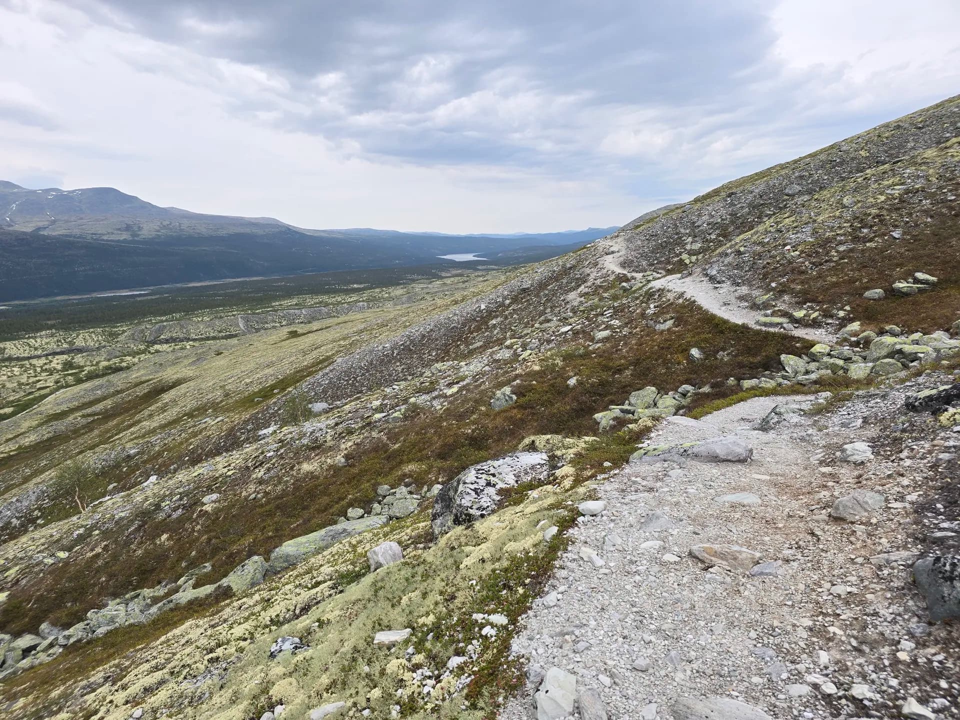 Hiking trail on a rocky alpine mountain slope with distant valley and lake under a cloudy sky.