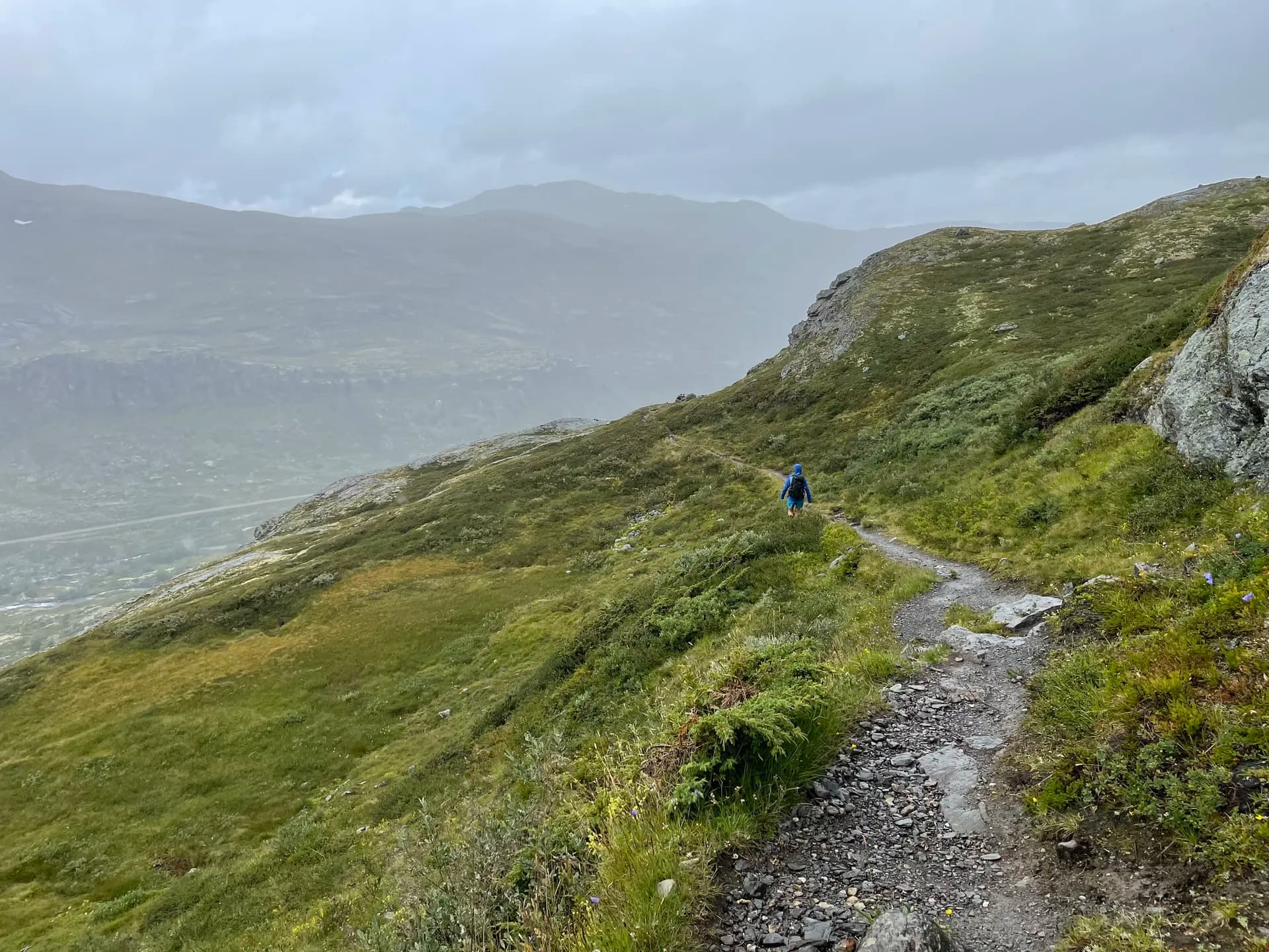 Hiker on a mountain trail in Aurlandsdalen, Norway, surrounded by green hills and misty peaks.