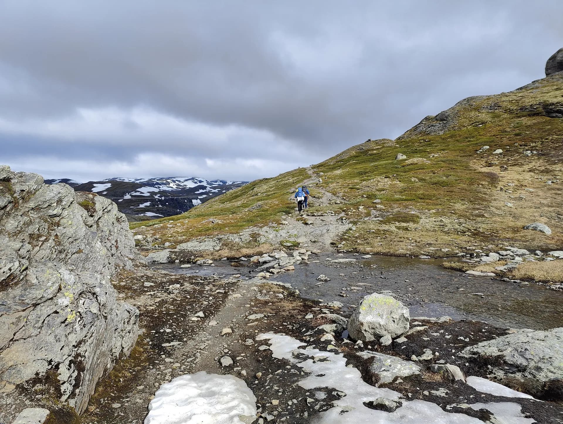Hikers on a rocky mountain trail in Aurlandsdalen, Norway, with snow-capped peaks and a stream.
