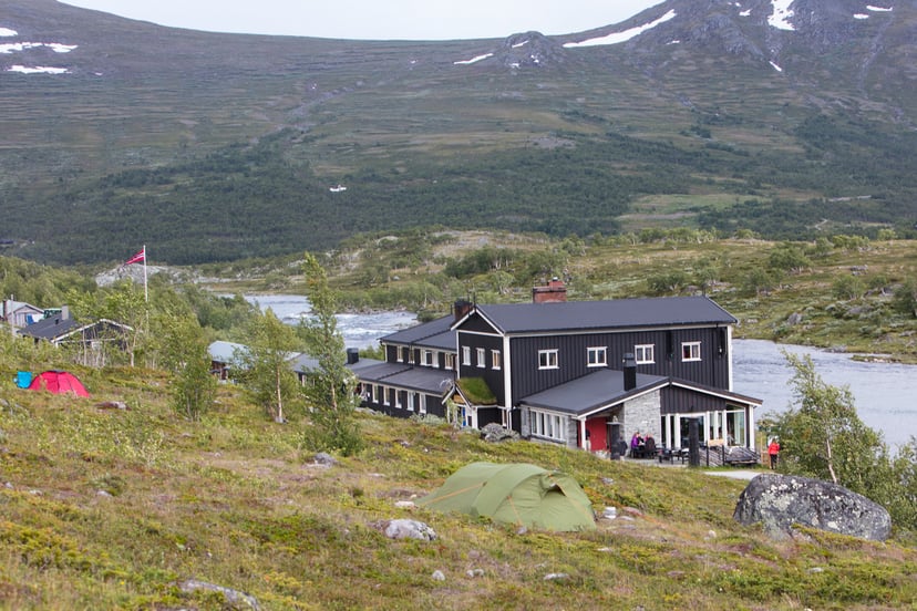 Camping near a black wooden mountain lodge by a river with snowy peaks in background