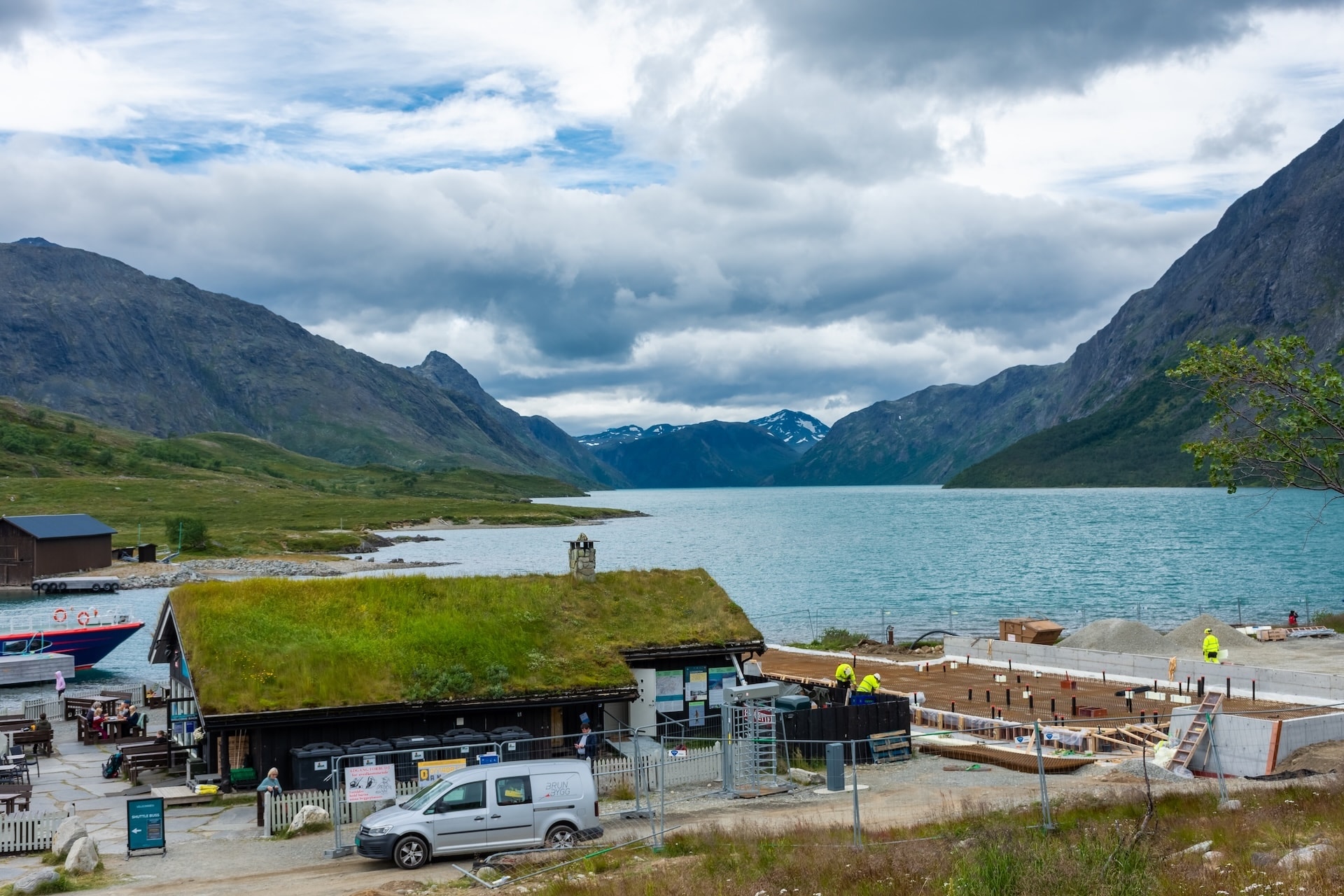 Building with grass roof by lake surrounded by mountains; construction underway.