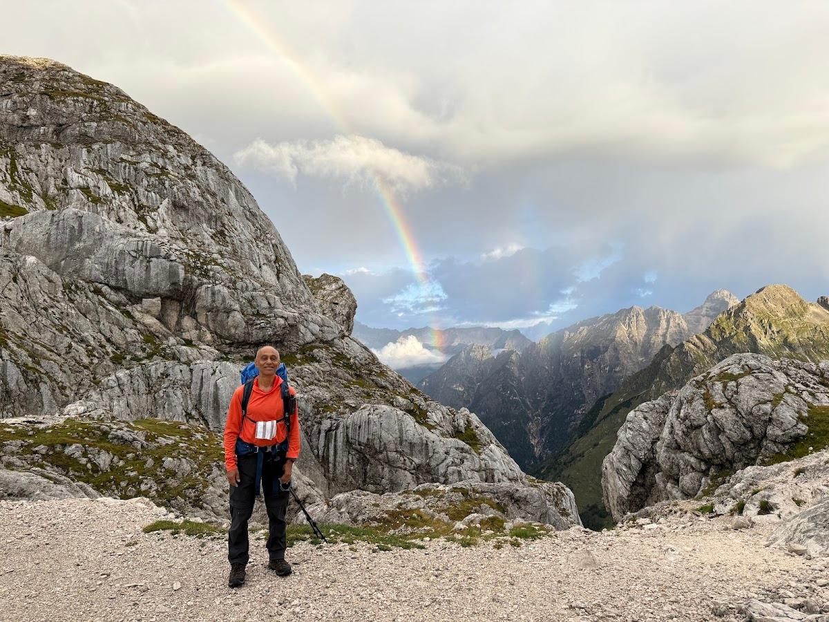 Hiker with backpack standing on rocky trail beneath rainbow over rugged mountains