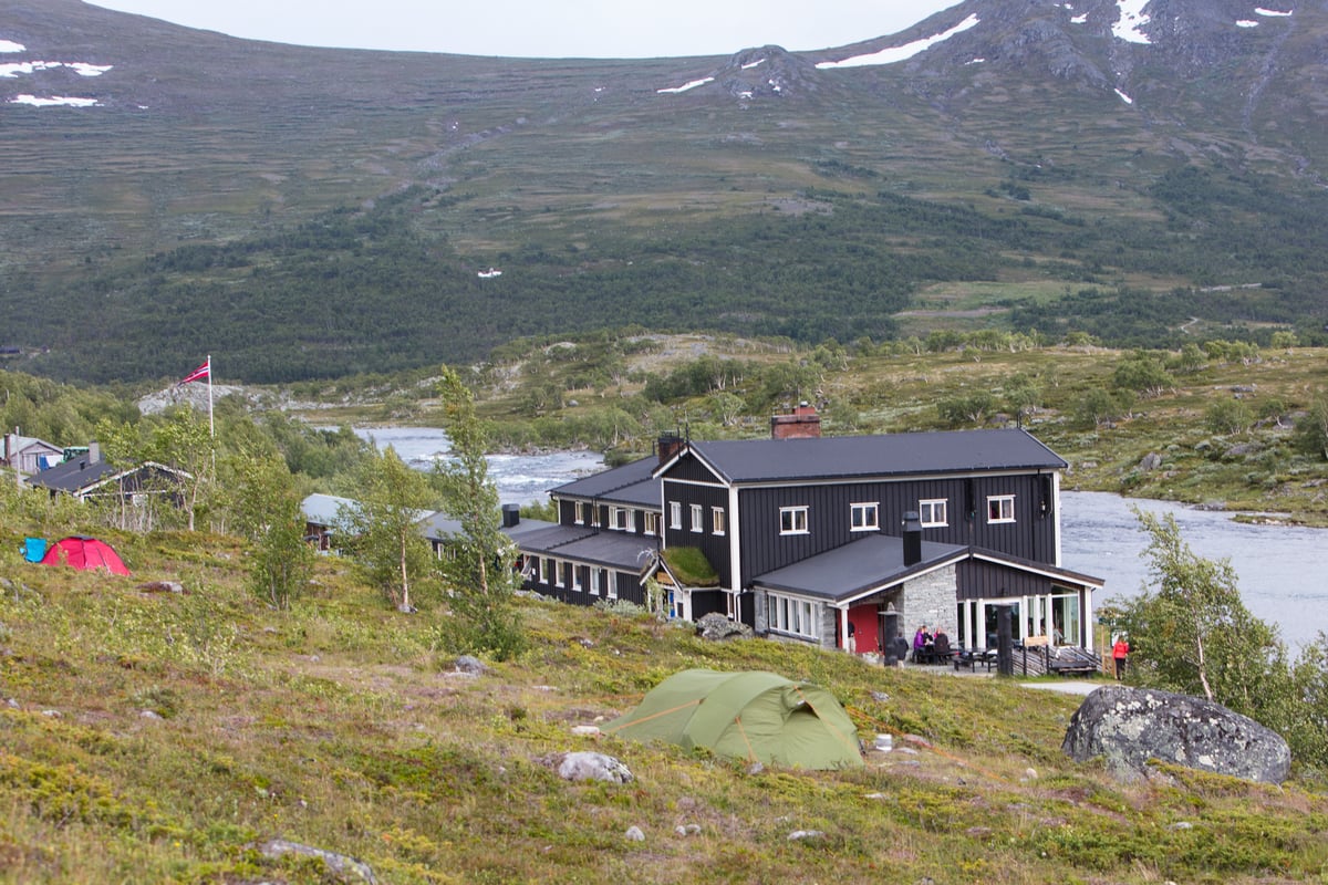 Camping near a black wooden mountain lodge by a river with snowy peaks in background