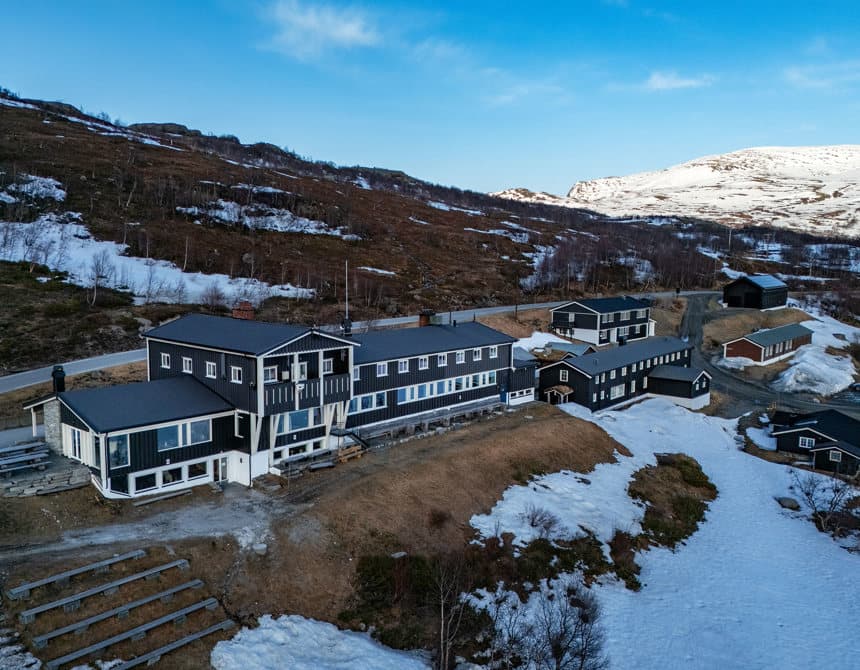 Black wooden buildings on a snowy mountainside with patches of brown grass under a blue sky.