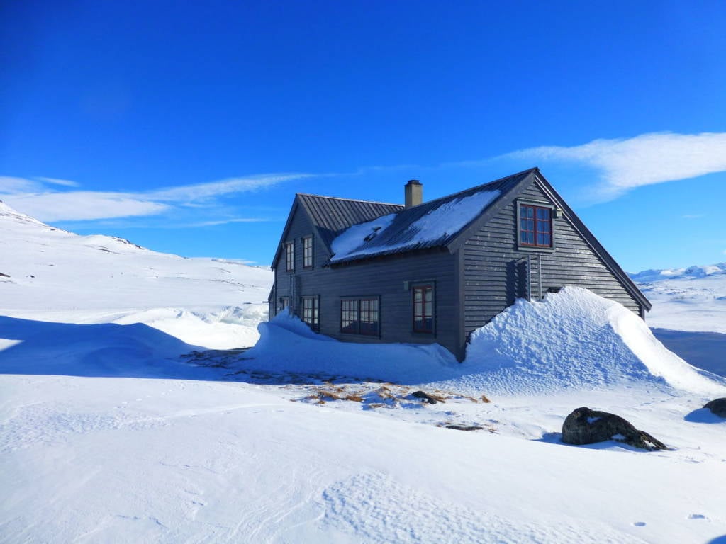 Wooden cabin buried in deep snow under a bright blue sky in a vast snowy landscape.