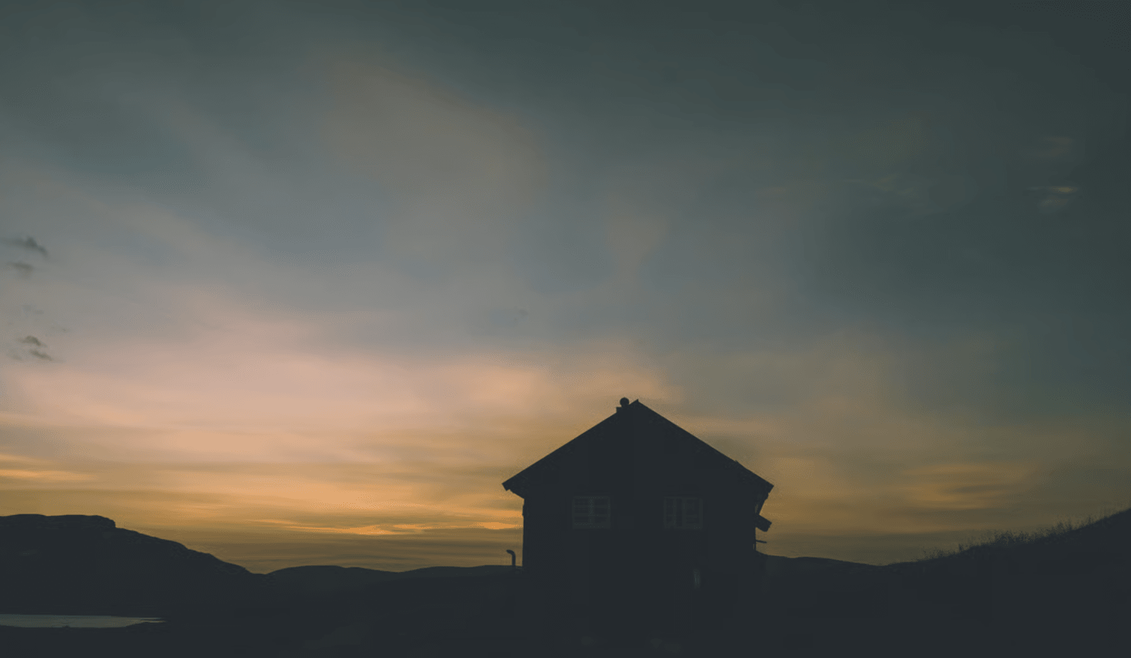 Silhouette of a house against a colorful sunset sky over rolling hills and water.