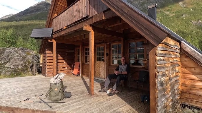 Hiker resting on porch of wooden cabin with backpack and trekking poles near mountains.