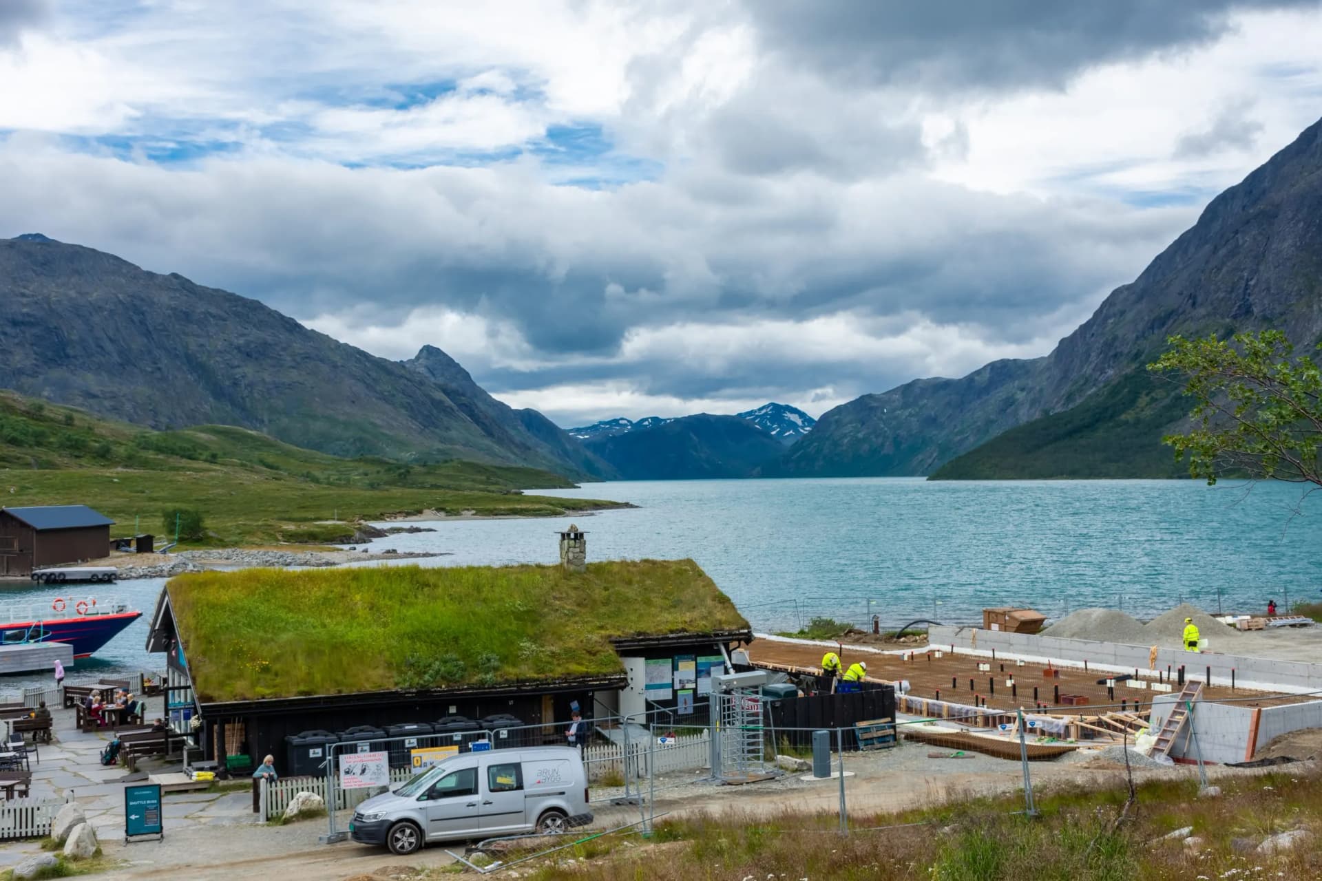 Gjendesheim Lodge with grass roof by lake, mountains, and construction site.