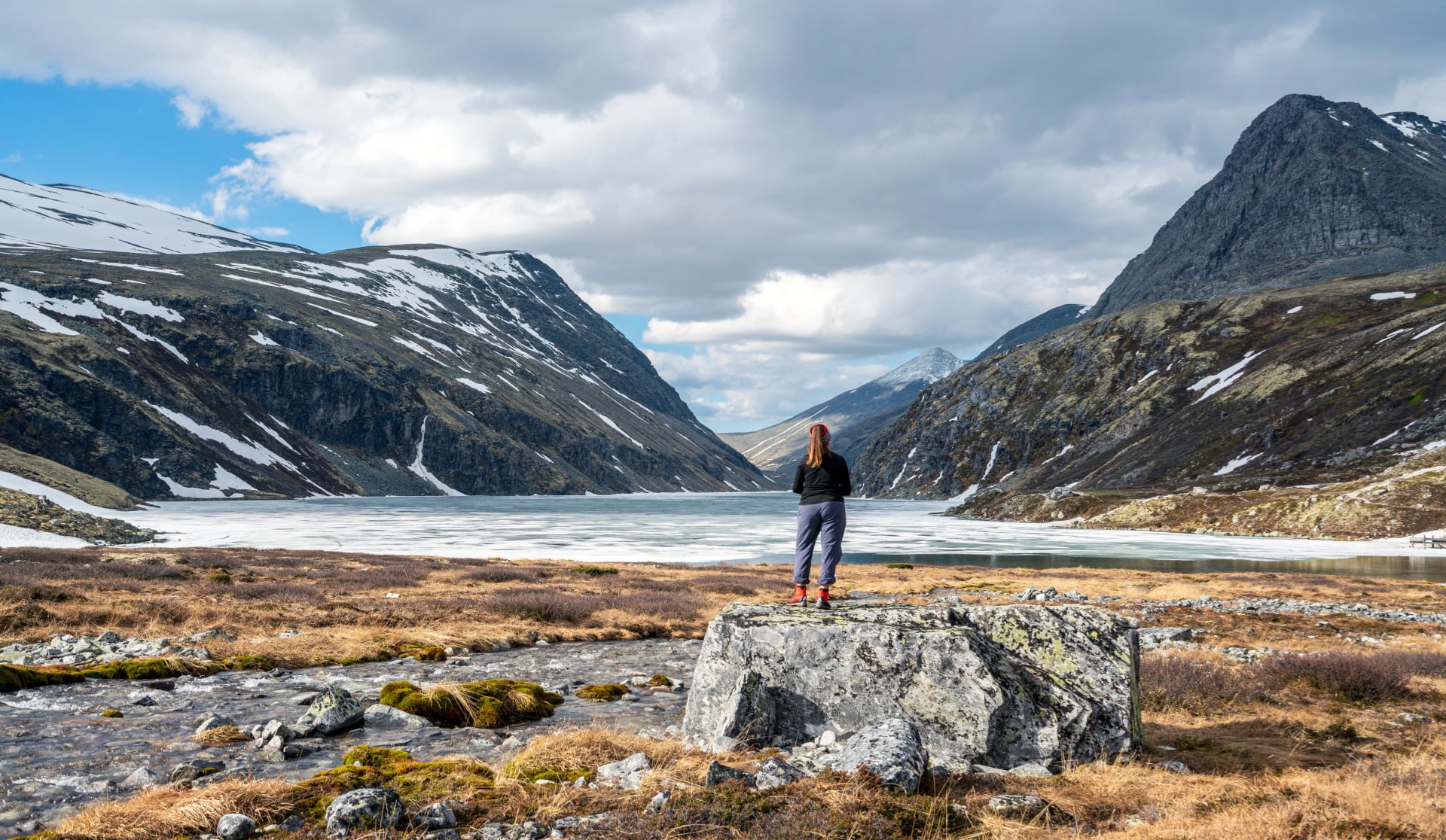Hiker woman standing on a rock looking towards icy lake and mountain scenery in Rondane national park in Norway.