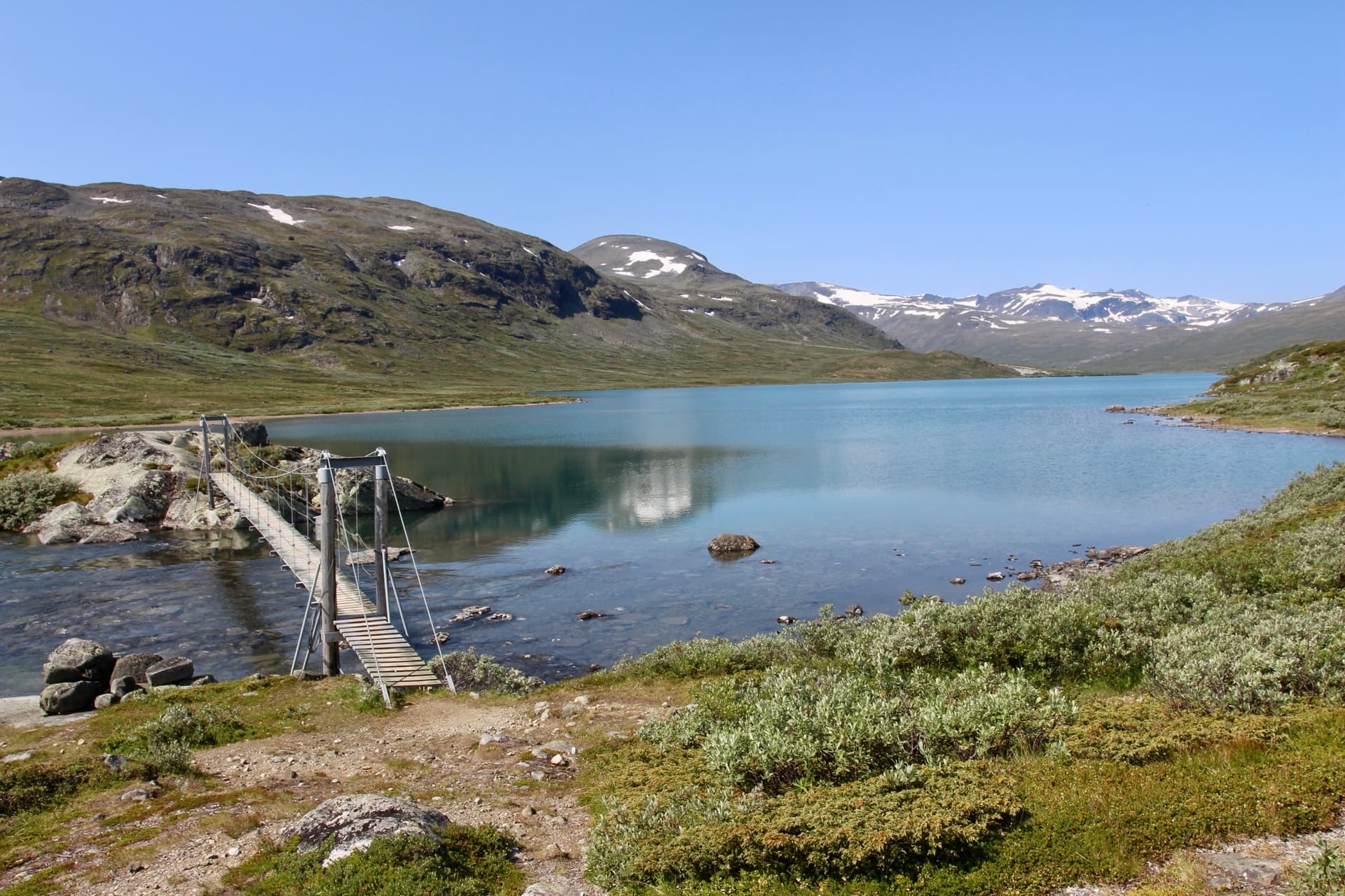 Footbridge crossing a stream by an alpine lake with snow-capped mountains