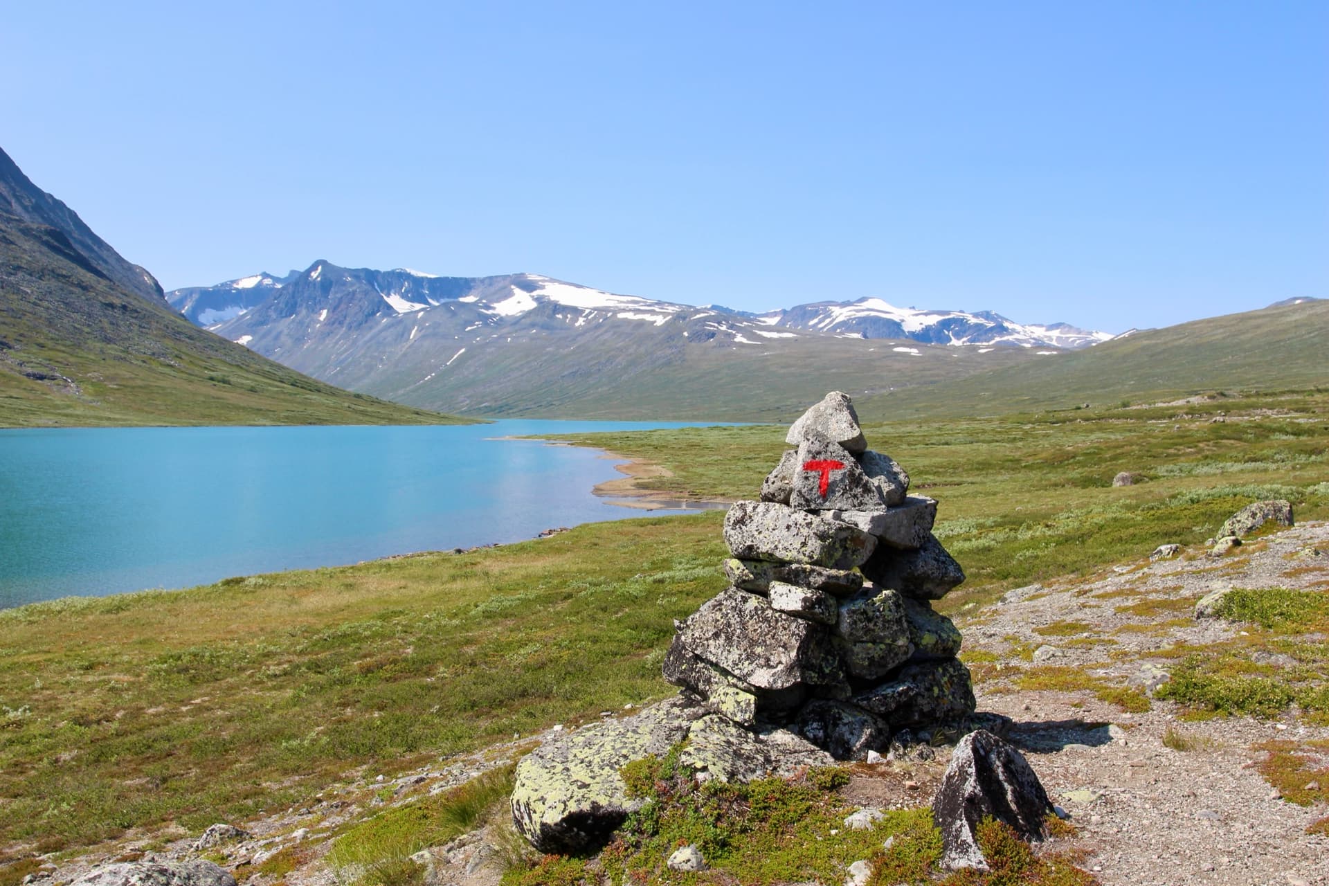 Cairn trail marker with red paint on stones in alpine mountain landscape with turquoise lake